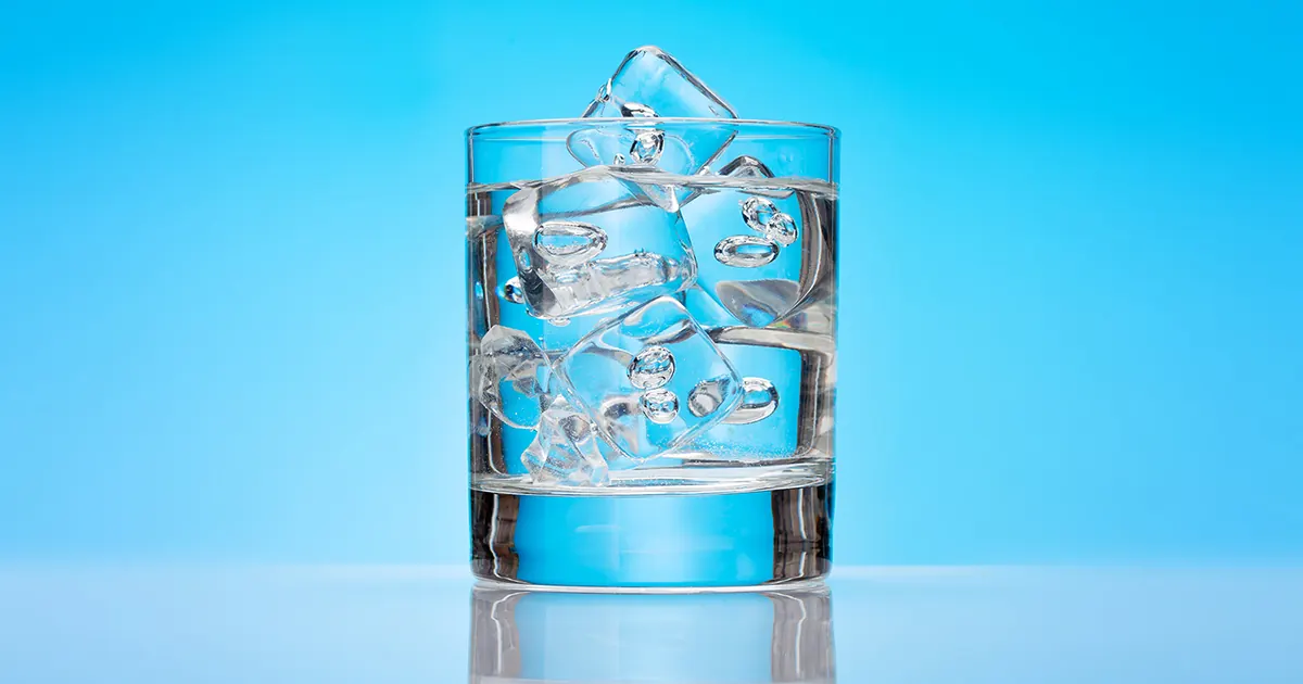 A glass tumbler of water and ice on a shiny surface, set against a twilight blue background