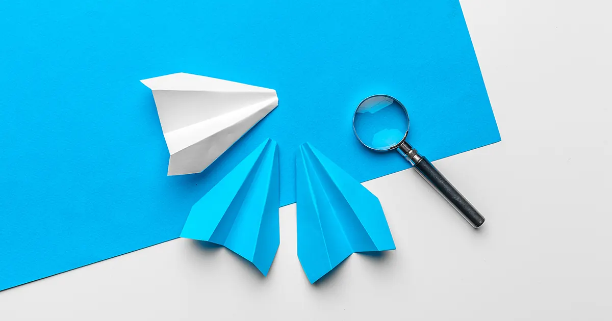 Paper planes (one white, two blue) and a magnifying glass arranged on a contrasted bright blue and white background