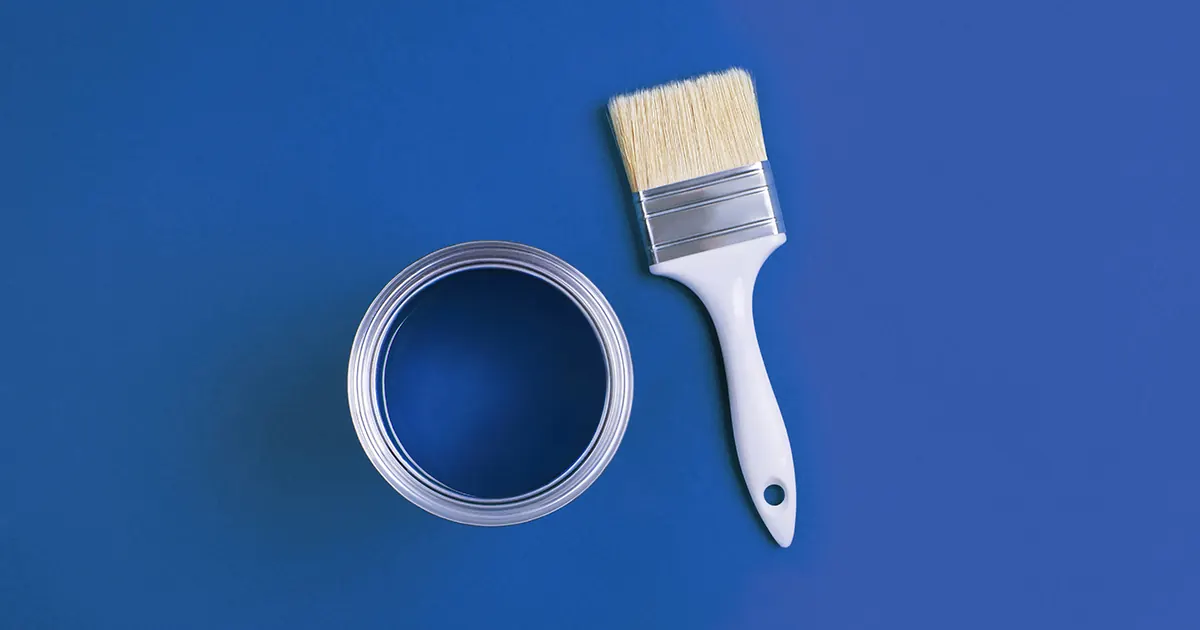 Plan view of an open tin of paint and a new paintbrush laid beside it on a medium blue background (the same shade of blue as the paint in the tin)